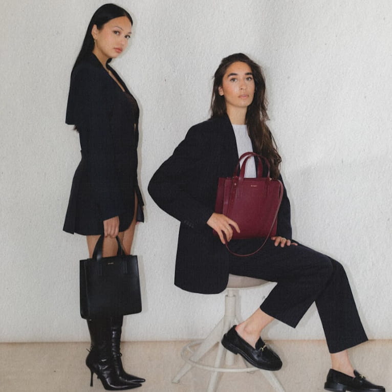 Two brunette Models wearing Blazers carrying the Iris Mare Jude Mini Tote bags in Midnight black and Lily Maroon